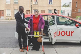 A passenger using a mobility aid exits a taxi with the help of the driver.